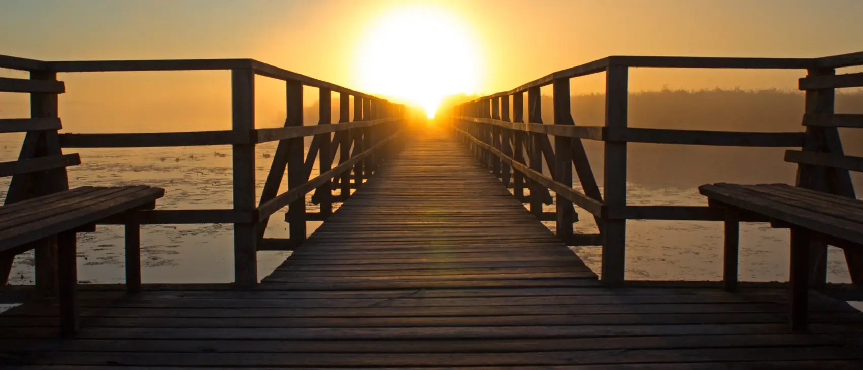 Pasarela de madera sobre un lago que apunta hacia el sol naciente